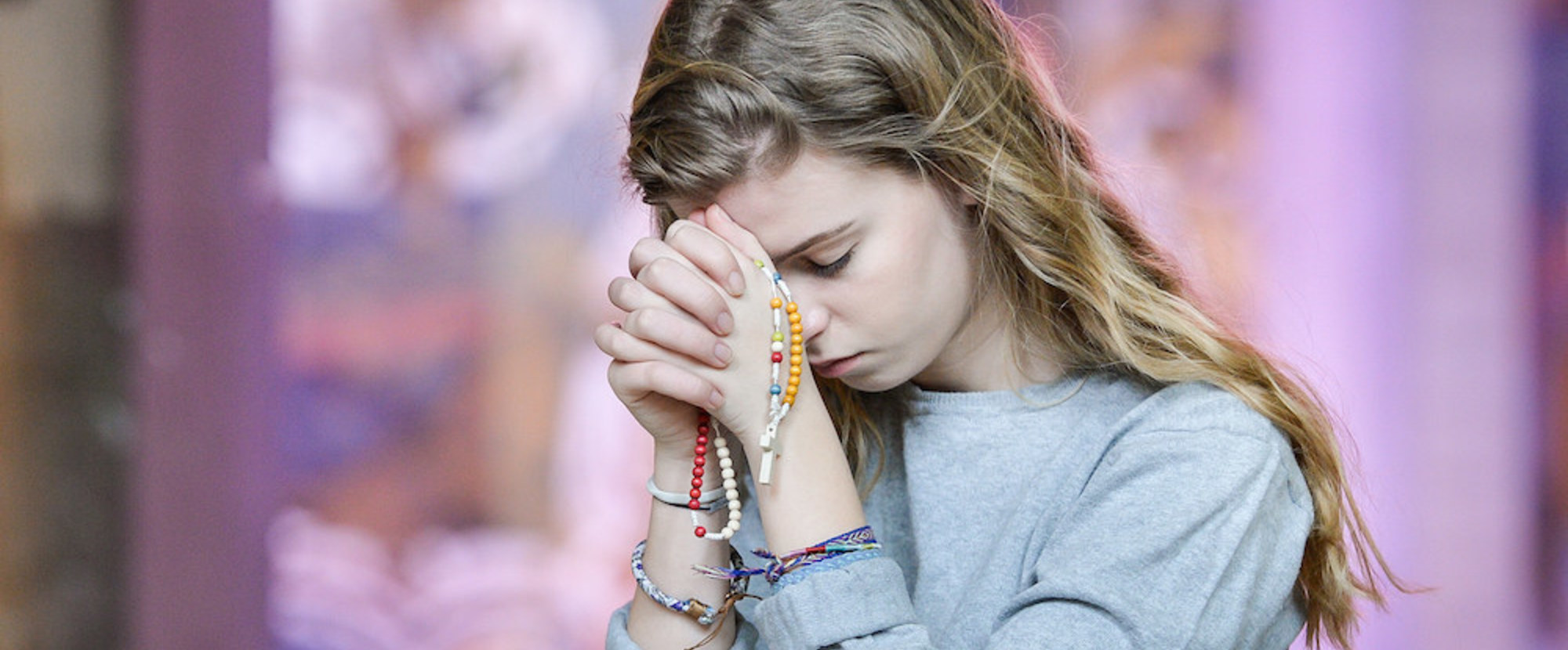 Eine junge, blonde Frau betet, ihren Kopf auf ihre gefalteten Hände gelegt, kniend in einer Kirchenbank am 27. Februar 2018. Ihre Augen sind geschlossen, in der Hand hält sie einen Rosenkranz, sie trägt bunte Armbänder aus Stoff. (Aufnahmeort unbekan