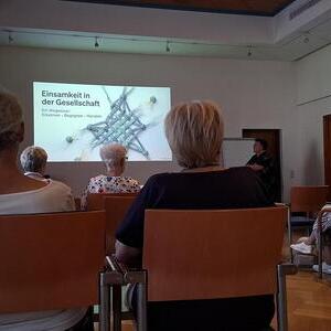 Menschen hören einem Vortrag zu und sehen eine Power Point Folie mit Titel des Vortrags