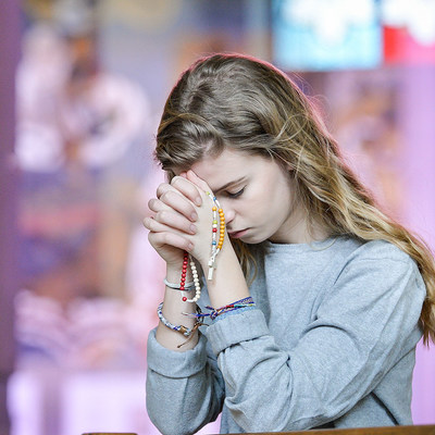 Eine junge, blonde Frau betet, ihren Kopf auf ihre gefalteten Hände gelegt, kniend in einer Kirchenbank am 27. Februar 2018. Ihre Augen sind geschlossen, in der Hand hält sie einen Rosenkranz, sie trägt bunte Armbänder aus Stoff. (Aufnahmeort unbekan