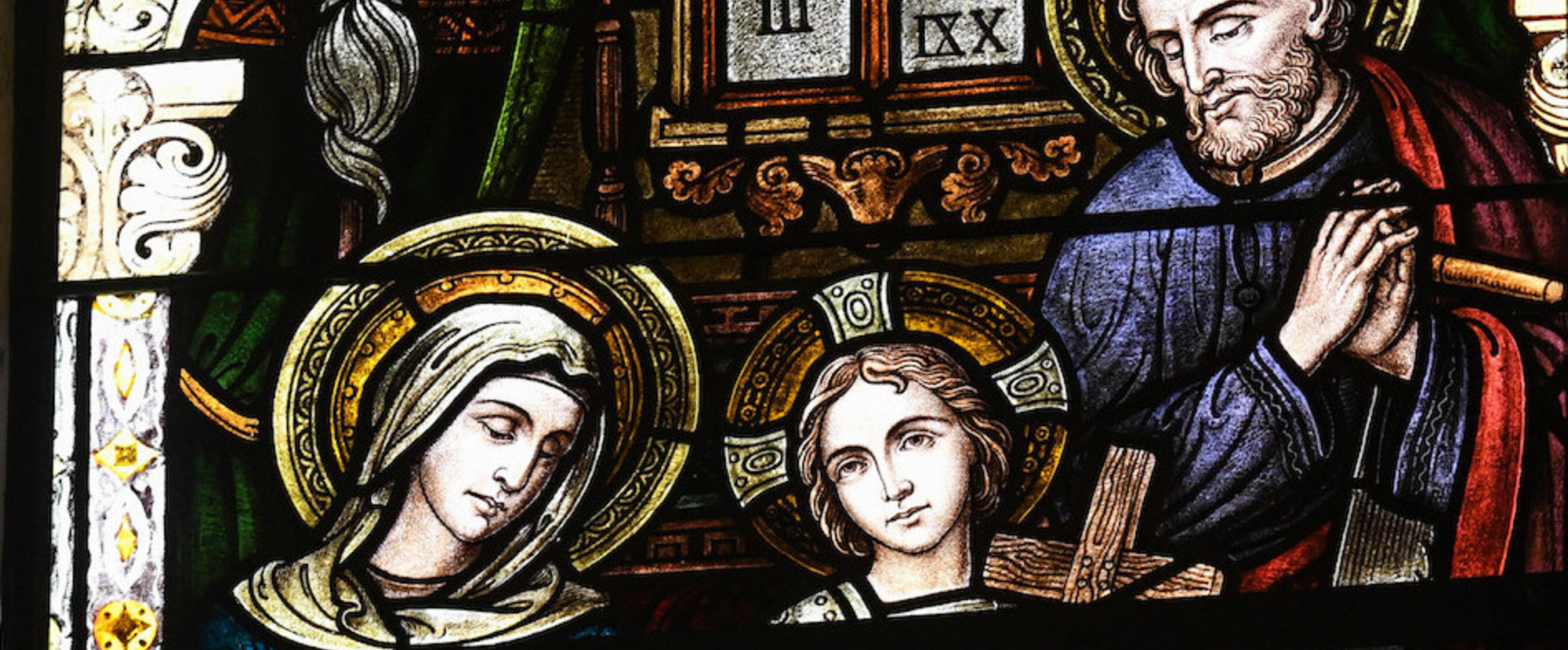 Kirchenfenster mit einer Darstellung der Heiligen Familie in der Kirche Sankt Rochus in Bonn am 12. Juli 2021. Maria hat den Kopf gesenkt, Josef steht an der Werkbank und Jesus hält ein Kreuz in den Händen.